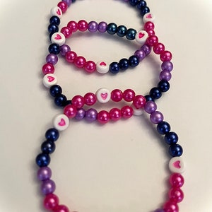 Pode incluir: Três pulseiras de contas em tons de rosa, roxo e azul com contas brancas com corações rosa.