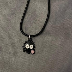 May include: Black pendant necklace featuring a cartoon soot sprite character with white eyes and a pink star. The pendant hangs from a black cord necklace. The background is a gray fabric.