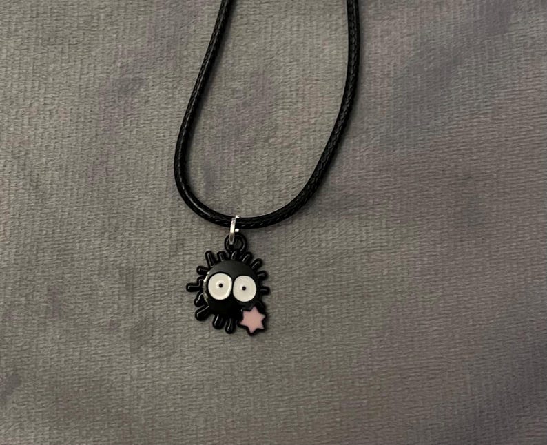 May include: Black pendant necklace featuring a cartoon soot sprite character with white eyes and a pink star. The pendant hangs from a black cord necklace. The background is a gray fabric.