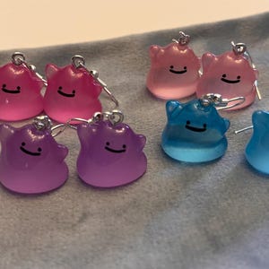 Può includere: Una collezione di orecchini fatti a mano in vari colori, tra cui rosa, viola e blu. Ogni orecchino presenta un design a forma di personaggio sorridente con un gancio color argento. Gli orecchini sono esposti su una superficie grigia.