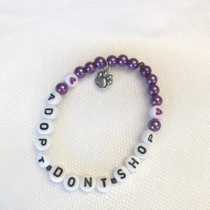 May include: Purple and white beaded bracelet with the words "I DON'T SHOP, ADOPT" and a small silver paw print charm.