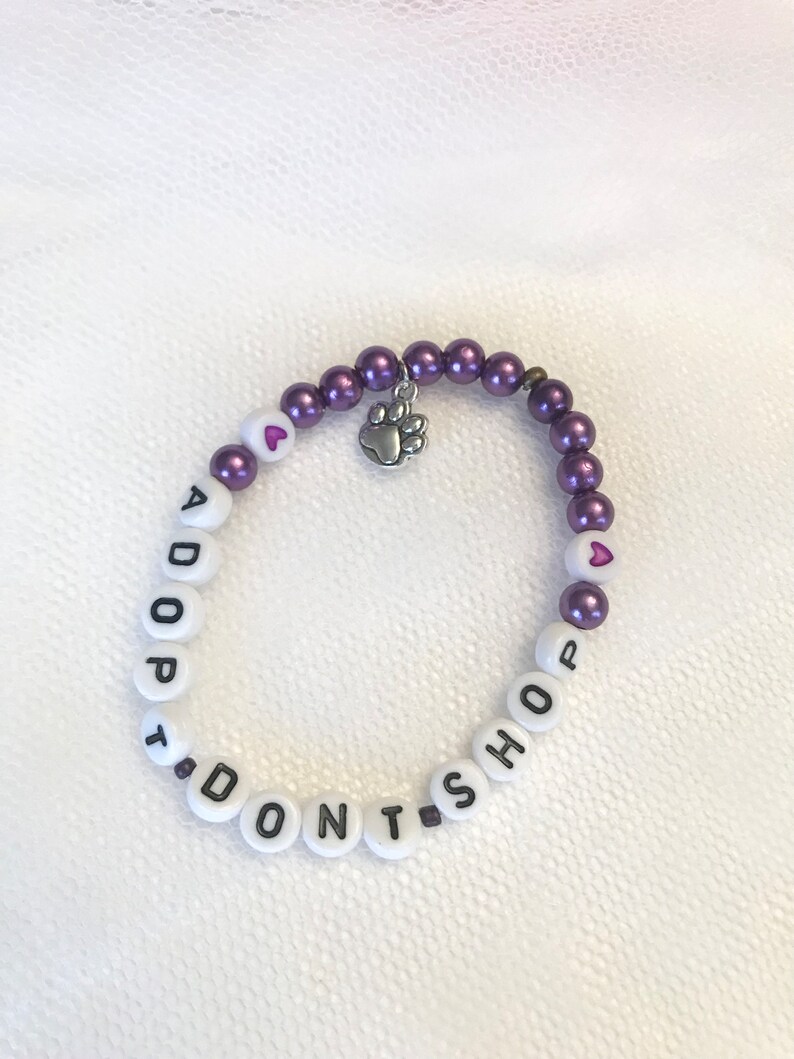 May include: Purple and white beaded bracelet with the words "I DON'T SHOP, ADOPT" and a small silver paw print charm.