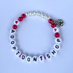 May include: White and red beaded bracelet with the words "Adopt Don't Shop" and a silver paw print charm.