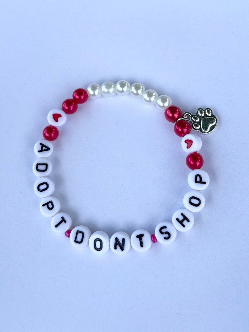 May include: White and red beaded bracelet with the words "Adopt Don't Shop" and a silver paw print charm.