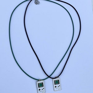 May include: Two necklaces with black and green cords and a silver clasp. Each necklace has a pendant shaped like a vintage handheld video game console with a green screen and white buttons.