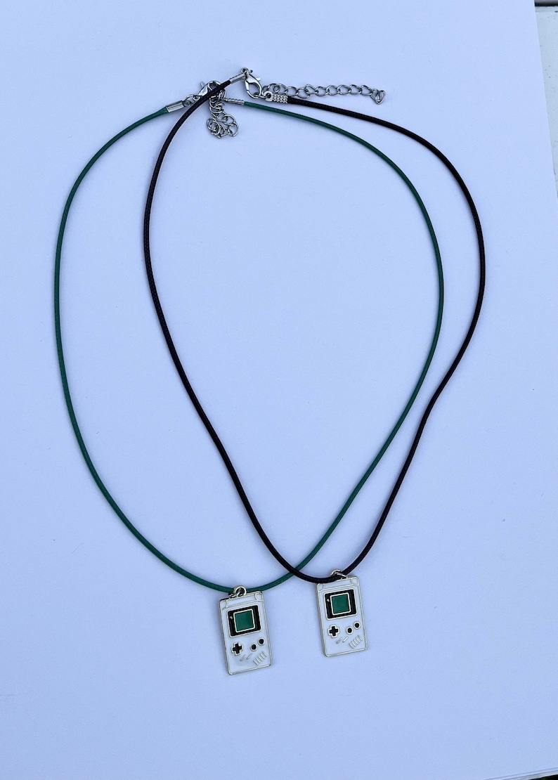 May include: Two necklaces with black and green cords and a silver clasp. Each necklace has a pendant shaped like a vintage handheld video game console with a green screen and white buttons.