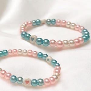 Op de afbeelding: Twee kralenarmbanden met afwisselende roze, witte en blauwe kralen. Elke armband heeft een witte hartbedel.