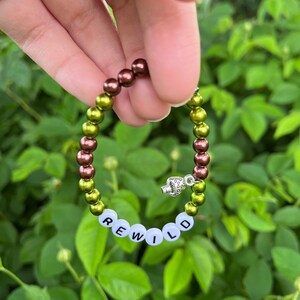 Pode incluir: Um bracelete de contas verdes e marrons com um fecho de prata em forma de cogumelo. O bracelete tem contas brancas com a palavra "REWILD" escrita.