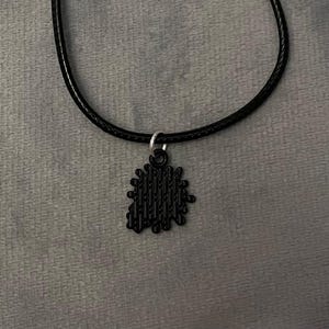 May include: Black pendant necklace. The necklace features a black, textured pendant suspended from a black cord. The pendant has a unique, irregular shape with vertical lines. The necklace is displayed against a gray background.