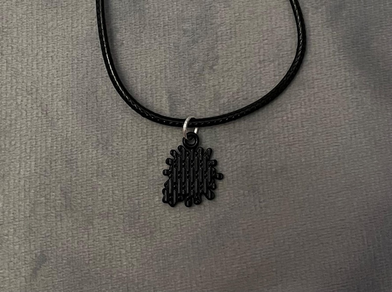 May include: Black pendant necklace. The necklace features a black, textured pendant suspended from a black cord. The pendant has a unique, irregular shape with vertical lines. The necklace is displayed against a gray background.