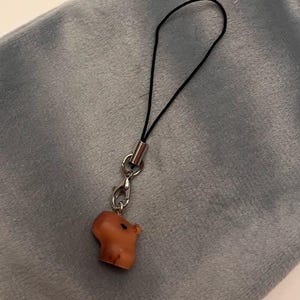 Puede incluir: Un pequeño amuleto de cerdo marrón con un cordón negro y herrajes metálicos plateados. El amuleto está unido a un lazo de cordón negro, adecuado para usar como llavero o amuleto de teléfono. El amuleto de cerdo es de color marrón claro.