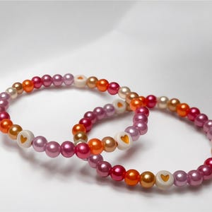 Könnte beinhalten: Zwei Perlenarmbänder mit einem Farbverlauf aus rosa, orange und goldenen Perlen. Jedes Armband hat eine weiße Perle mit einem goldenen Herzdesign.