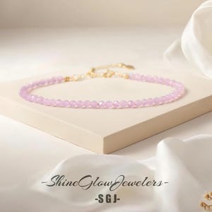 May include: A delicate, light purple beaded necklace with a gold-colored clasp and chain. The necklace is displayed on a small, off-white platform, with a white satin fabric in the background. The text "-Shine Glow Jewelers- -SGJ-" is visible.