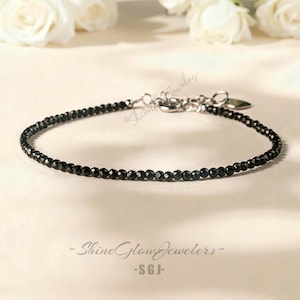May include: A fine black beaded bracelet with a silver clasp. The bracelet is made up of small, round, black beads. The background features white roses and the text "- Shine Glow Jewelers - SGI -."