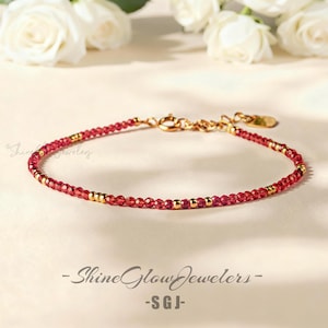 May include: A delicate bracelet featuring small, red beads interspersed with gold-colored accents. The bracelet is secured with a gold clasp and is displayed on a light-colored surface. The text "Shine Glow Jewelers - SGJ-" is visible.