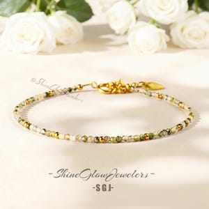 May include: Delicate beaded bracelet featuring small, round, green and clear gemstones with gold accents. The bracelet has a gold clasp and a small gold charm. The text "Shine Glow Jewelers - SGJ" is at the bottom.