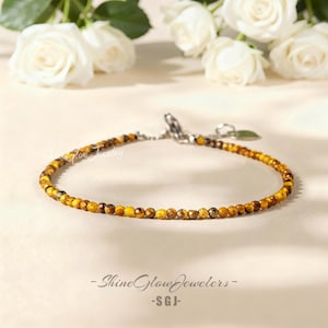 May include: A delicate bracelet featuring small, round, golden-brown beads. The bracelet has a silver-colored clasp and a small charm. The background includes white roses and the text "Shine Glow Jewelers - SGJ".