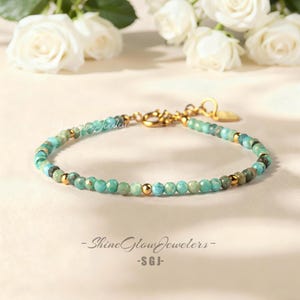 May include: A turquoise beaded bracelet with gold detailing. The bracelet is made up of small, round turquoise beads, gold-coloured beads, and a gold clasp. White roses are in the background, with the text "Shine Glow Jewelers - SGJ".