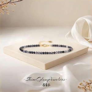 May include: A delicate silver bracelet with small, dark blue and white beads. The bracelet is displayed on a white surface with a white fabric background.