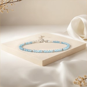 May include: A delicate silver chain necklace with light blue gemstone beads. The necklace is displayed on a white surface.