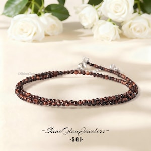 May include: A delicate, layered necklace featuring small, faceted, reddish-brown beads. The necklace has a silver clasp and is displayed on a light background with white roses. The text "Shine Glow Jewelers - SGJ-" is at the bottom.