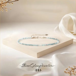 May include: A delicate silver chain necklace with light blue gemstone beads. The necklace is displayed on a white surface with a white fabric background.