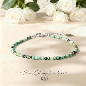 May include: A delicate beaded bracelet featuring small, round turquoise and black beads strung on a silver chain. The bracelet is displayed with white roses in the background. The text "- Shine Glow Jewelers -" and "- SGJ -" is visible.
