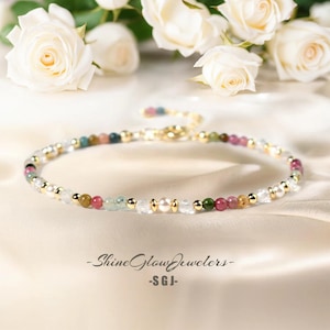 Könnte beinhalten: Eine zarte goldene Kettenkette mit einer Vielzahl von bunten Edelsteinen, darunter rosa, grün und blau, durchsetzt mit weißen Perlen und klaren Kristallen. Die Kette wird auf einem weißen Satin-Hintergrund präsentiert. -ShineGlowJewelers- -SGJ-