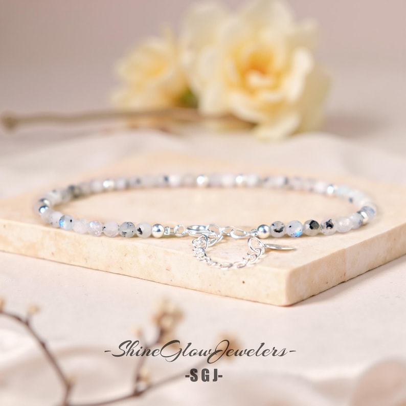 Op de afbeelding: Een delicate zilveren kettingarmband met kleine witte en grijze edelsteen kralen. De armband heeft een karabijn sluiting en een klein zilveren bedeltje.