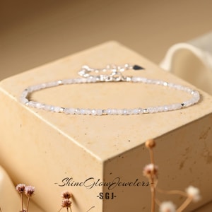 Puede incluir: Un delicado brazalete de plata con pequeñas cuentas blancas. El brazalete está sobre una superficie marrón claro con un fondo floral.