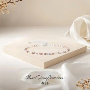 May include: Delicate beaded bracelet featuring small, colorful gemstones and silver-toned beads. The bracelet is displayed on a neutral-toned surface with a white ribbon in the background. The text "Shine Glow Jewelers - SGJ-" is visible.