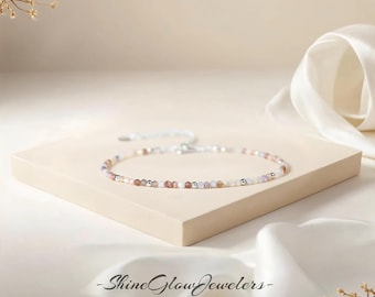 May include: Delicate beaded bracelet featuring small, colorful gemstones and silver-toned beads. The bracelet is displayed on a neutral-toned surface with a white ribbon in the background. The text "Shine Glow Jewelers - SGJ-" is visible.