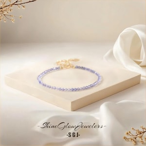 May include: A delicate bracelet made of small, light purple beads with a gold clasp. The bracelet is displayed on a white surface.