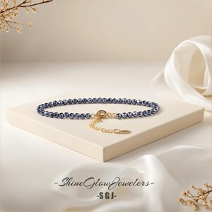 May include: A delicate blue crystal beaded bracelet with a gold chain and clasp. The bracelet is on a white surface with a white fabric background.