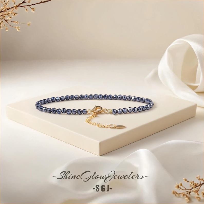 May include: A delicate blue crystal beaded bracelet with a gold chain and clasp. The bracelet is on a white surface with a white fabric background.