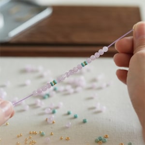 May include: A string of pale purple beads interspersed with turquoise and gold-coloured accents. The beads are threaded on a thin, light purple cord. Small beads in various colours are scattered around, suggesting jewellery making or crafting.
