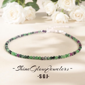 May include: A necklace with small, faceted beads in green, purple, and grey. The necklace is strung on a silver chain. The background is a soft, neutral colour with white roses. The text "- Shine Glow Jewelers -" and "- SGJ -" is at the bottom.