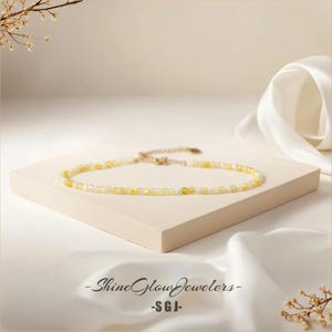May include: A delicate gold necklace with small, white and yellow beads. The necklace is displayed on a white surface with a white fabric background.