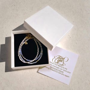 May include: A white box with a black interior containing a layered necklace with white and blue beads. The box also includes a thank you card with the text "Thank you. Your support means the world to me. This package can store your memories forever. Looking forward to see you again."
