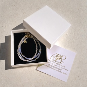 May include: A white gift box with a black interior, containing a triple-strand beaded bracelet with light blue and white beads. The box is open and a thank you card is visible with the text "Your support means the world to me. This package can store your memories forever. Looking forward to see you again."