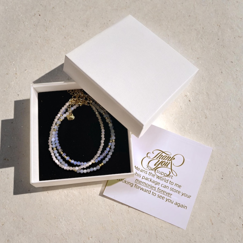 May include: A white gift box with a black interior, containing a triple-strand beaded bracelet with light blue and white beads. The box is open and a thank you card is visible with the text "Your support means the world to me. This package can store your memories forever. Looking forward to see you again."