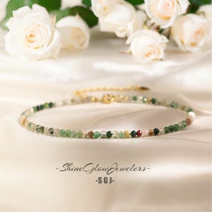 May include: A delicate gold chain necklace with small, multi-colored gemstone beads. The beads are a mix of green, brown, and white, with a few red beads scattered throughout. The necklace is displayed on a white satin background.