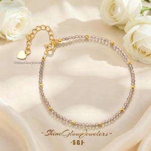 May include: A delicate bracelet featuring small, translucent, light gray faceted beads interspersed with tiny, polished gold-tone metal spheres. The piece includes a gold-tone chain extender with a heart-shaped charm and a spring ring clasp.