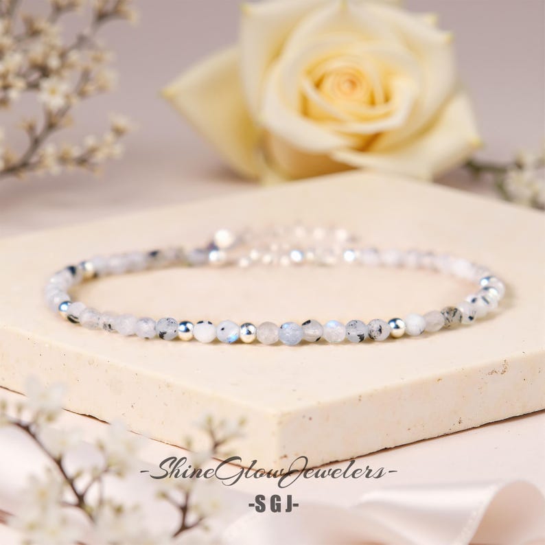 Op de afbeelding: Een delicate zilveren armband met witte maansteen kralen en zilveren tussenkralen. De armband ligt op een lichtbeige oppervlak.