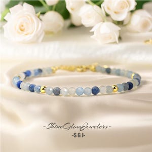 Könnte beinhalten: Ein zartes Armband aus blauen und weißen Edelsteinperlen mit goldenen Akzenten. Das Armband liegt auf einem weißen Stoffuntergrund. -ShineGlowJewelers- -SGJ-