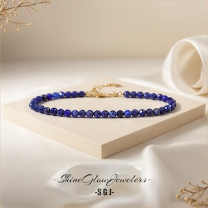 May include: A delicate necklace made of small, round, dark blue lapis lazuli beads. The necklace is on a white surface with a white fabric background.