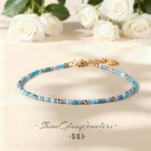 May include: Delicate beaded bracelet featuring small, round, blue and gold beads. The bracelet has a gold clasp and a small gold charm. The text "- Shine Glow Jewelers -" and "- SGJ -" is visible.