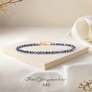 Könnte beinhalten: Eine zarte Halskette aus kleinen, dunkelblauen facettierten Perlen. Die Halskette wird auf einer weißen Oberfläche mit einem weißen Stoffhintergrund präsentiert. Der Text "Shine Glow Jewelers -SGJ- " ist am unteren Rand des Bildes sichtbar.