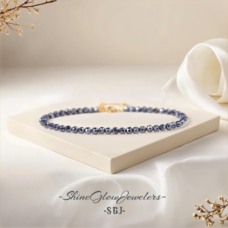 May include: A delicate necklace made of small, dark blue faceted beads. The necklace is displayed on a white surface with a white fabric background. The text "Shine Glow Jewelers -SGJ- " is visible at the bottom of the image.