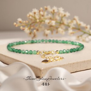 May include: A delicate green gemstone beaded bracelet with a gold heart charm and a gold clasp. The bracelet is on a white surface.